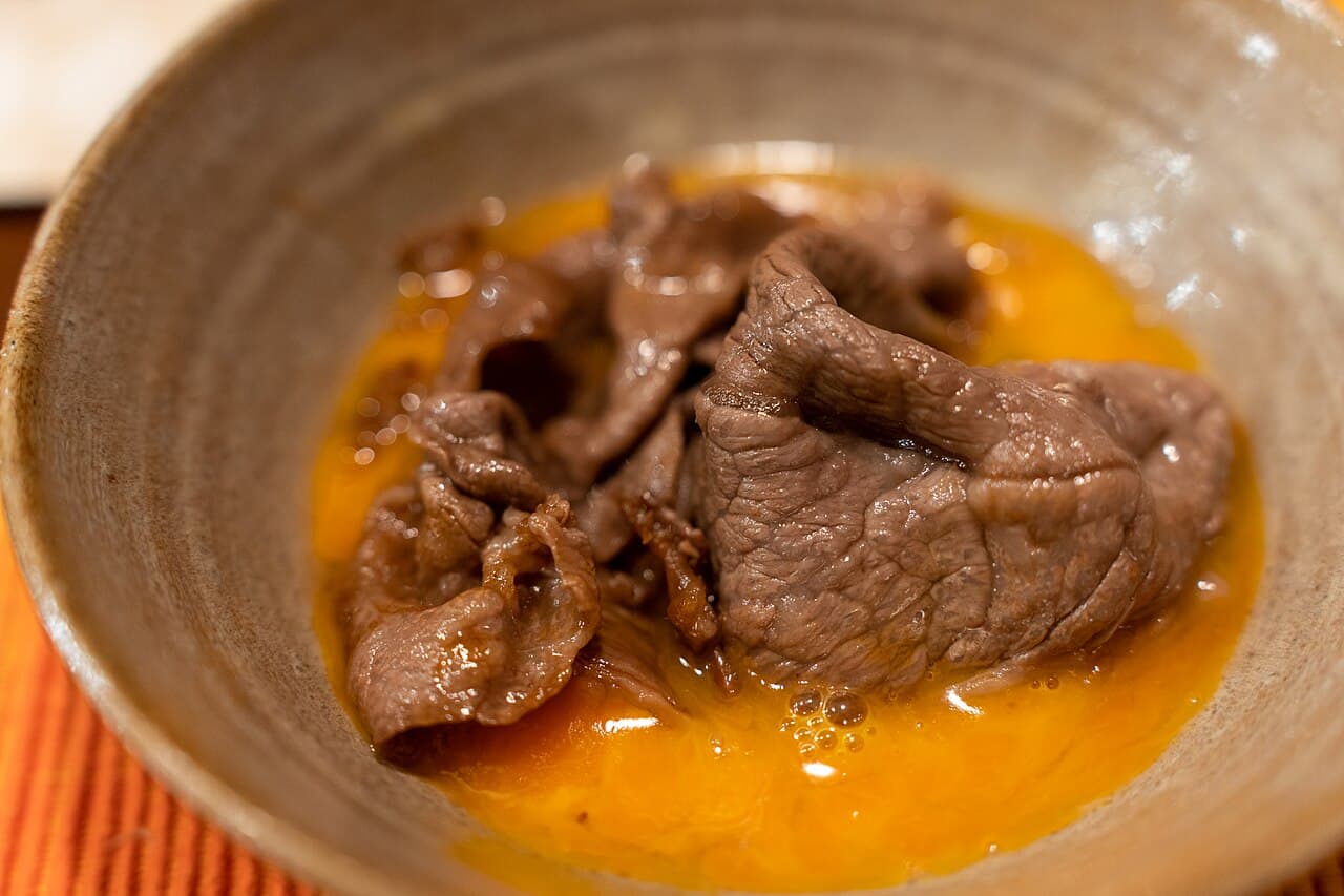 陶器の器の中で、溶いた生卵にくぐらせた、甘辛く煮えた薄切りの牛肉
