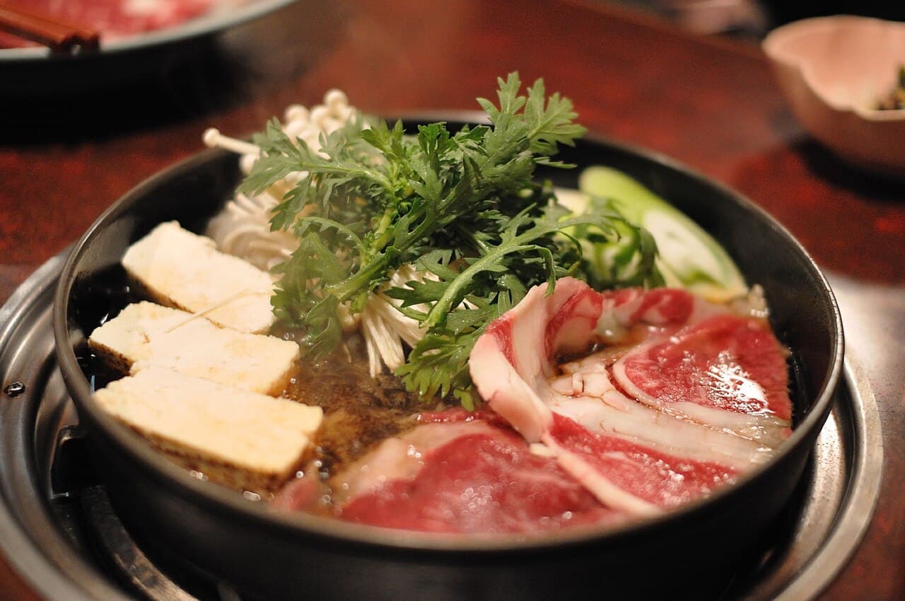 黒い鉄鍋に、霜降りの牛肉、焼き豆腐、春菊、えのきが並べられたすき焼き
