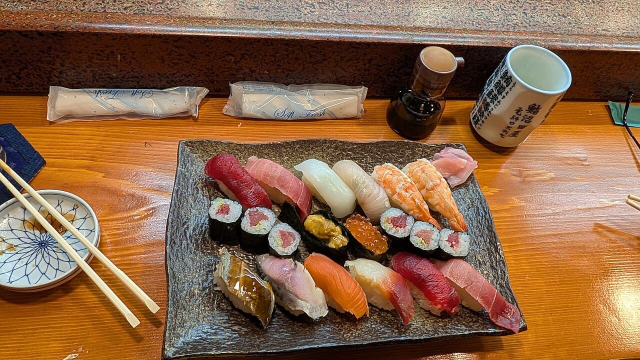 Nigiri sushi such as tuna and shrimp, with thin rolls, on a black square plate on a wooden counter, with a tea cup and a soy sauce bottle behind