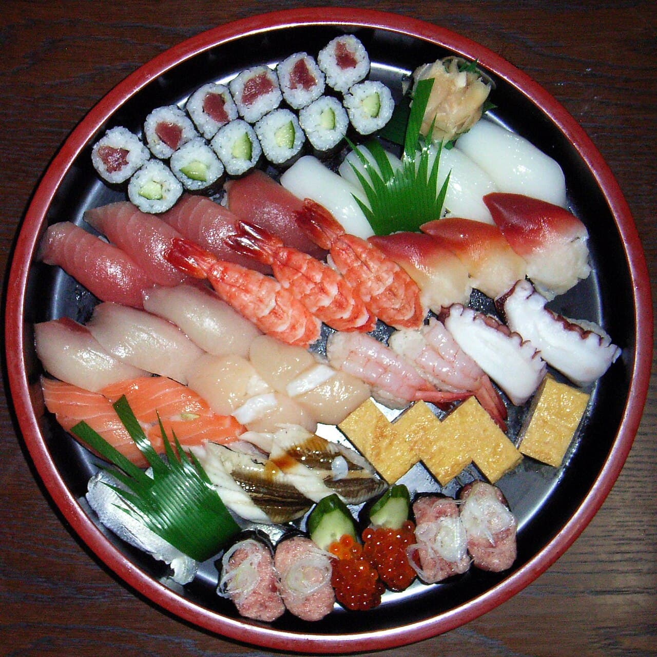 A round red and black sushi tub filled with tuna, shrimp, octopus, salmon roe, rolled omelet, thin rolls and more