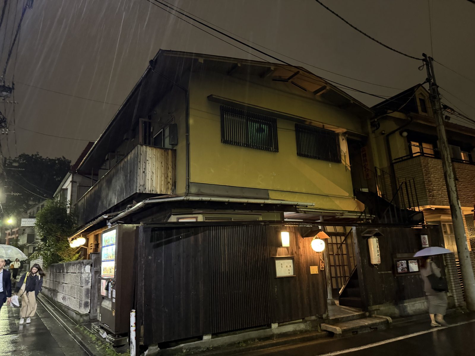 A rainy night in Kagurazaka — a quiet wooden building at a back-street corner, lit by hanging lanterns.